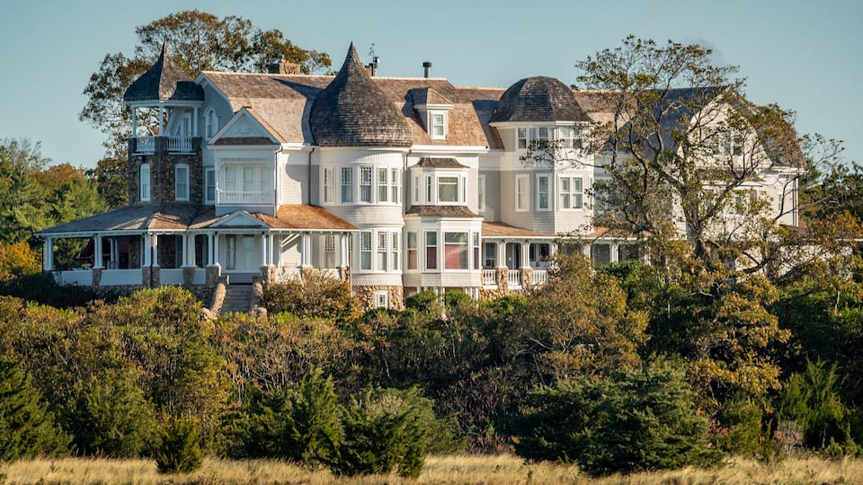 A large, multi-story house with a wrap-around porch, surrounded by dense trees and a grassy field.