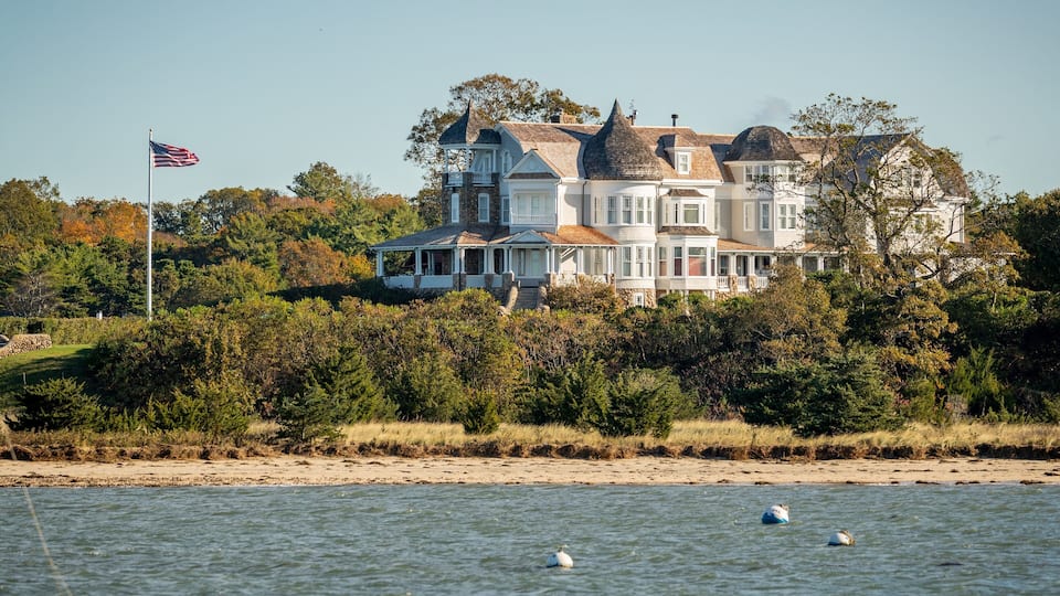 A large waterfront house with a flag, surrounded by trees and a sandy beach.