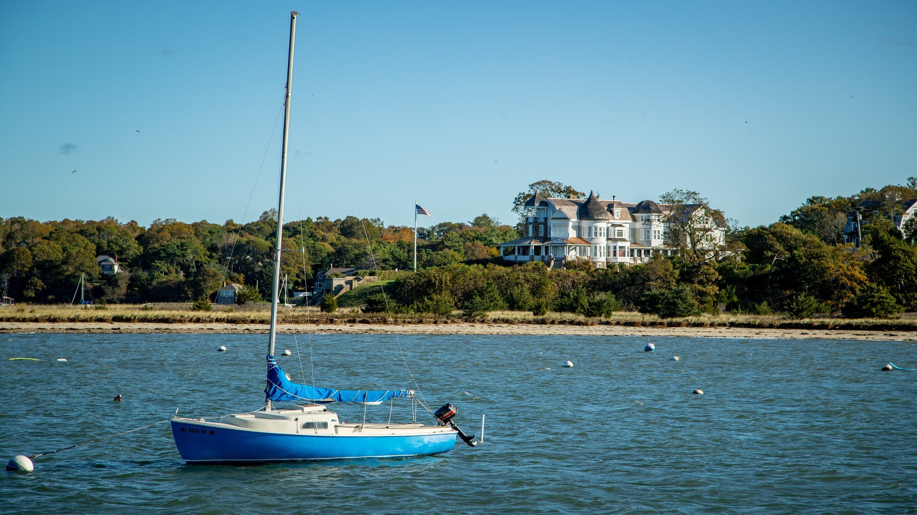 A sailboat on calm water with a large house in the background.