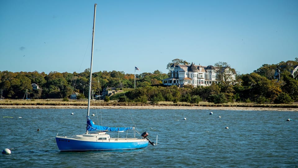 A sailboat on calm water with a large house in the background.