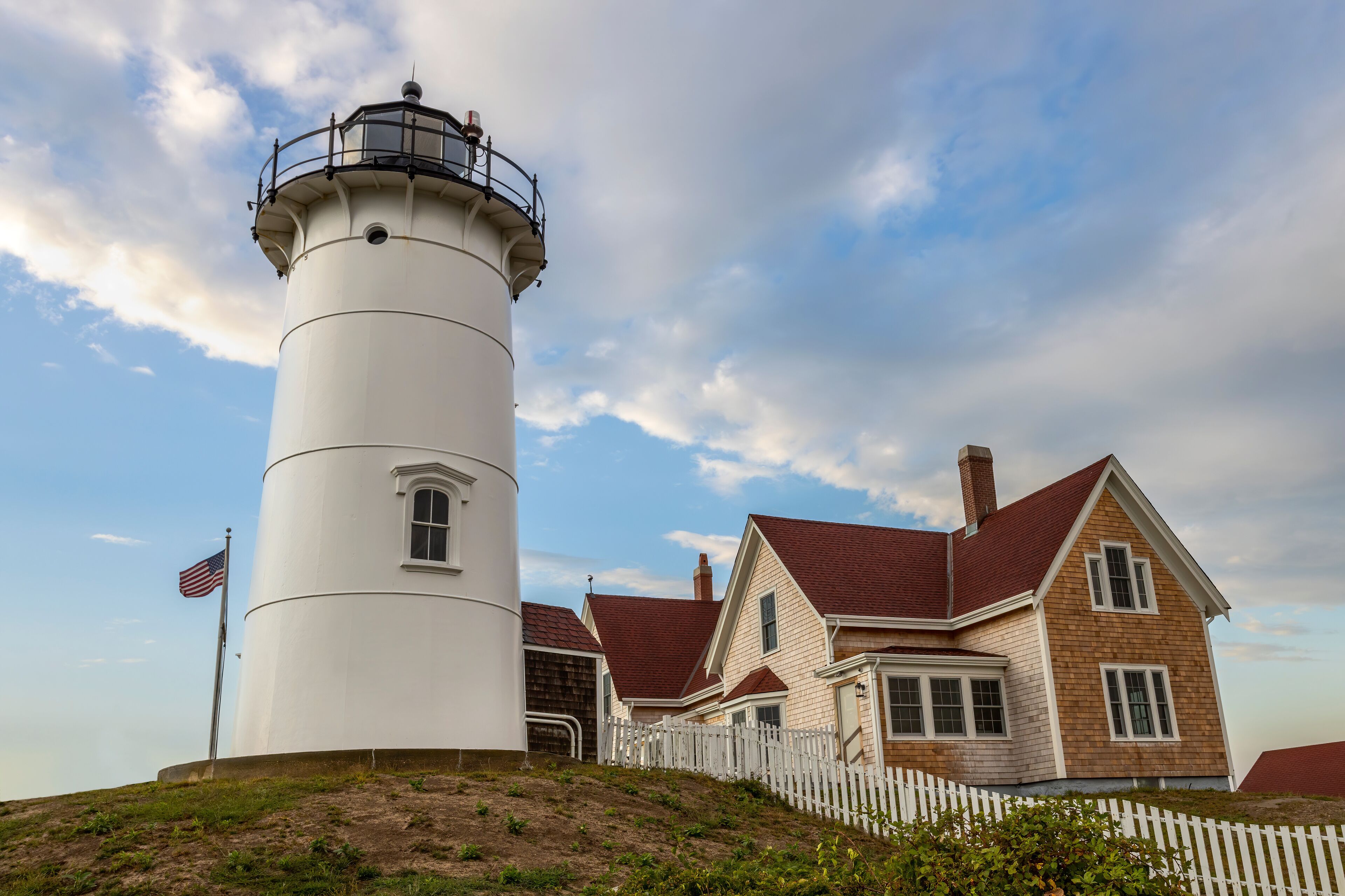 A white lighthouse with a red roof, a white picket fence, and a house with a red roof.