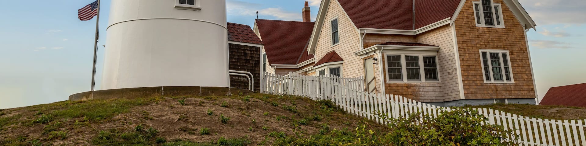 A white lighthouse with a red roof, a white picket fence, and a house with a red roof.