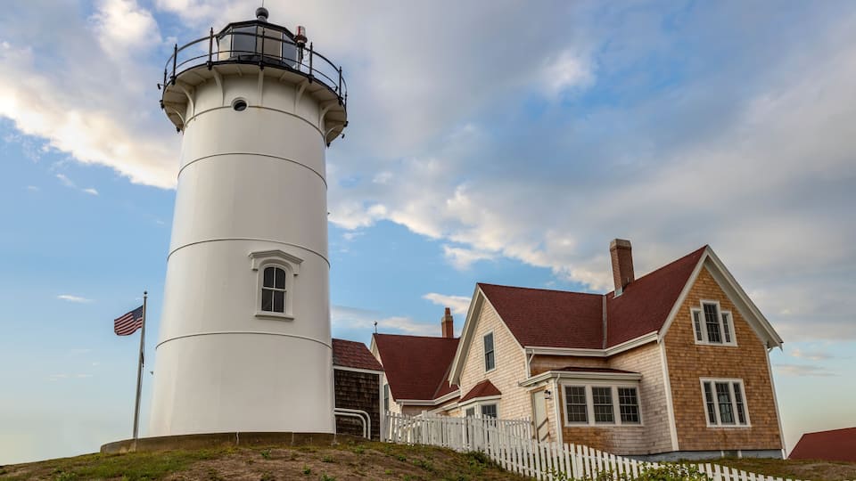 A white lighthouse with a red roof, a white picket fence, and a house with a red roof.