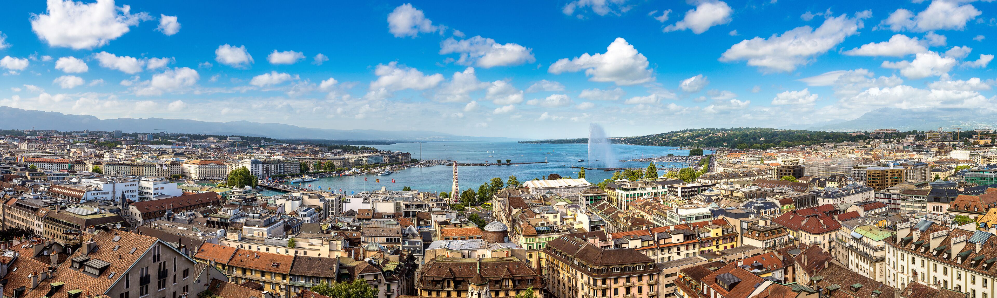 A panoramic view of a city with a river, mountains in the background, and a prominent fountain.