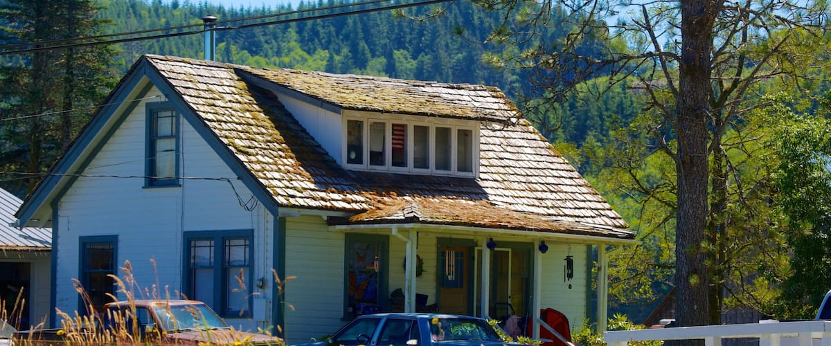 A rustic two-story house with a weathered roof, surrounded by tall trees and a white picket fence.