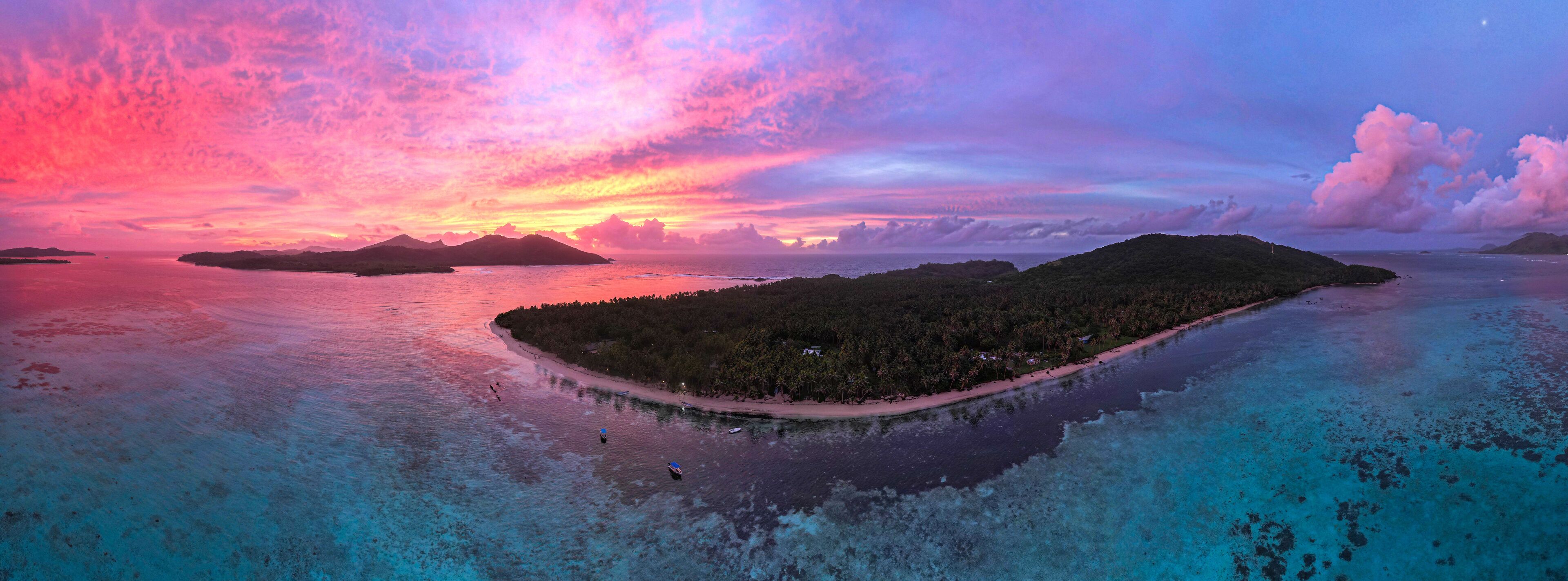 An aerial view of a tropical island surrounded by turquoise waters, with a pink and purple sunset in the background.