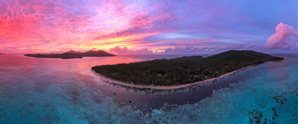 An aerial view of a tropical island surrounded by turquoise waters, with a pink and purple sunset in the background.