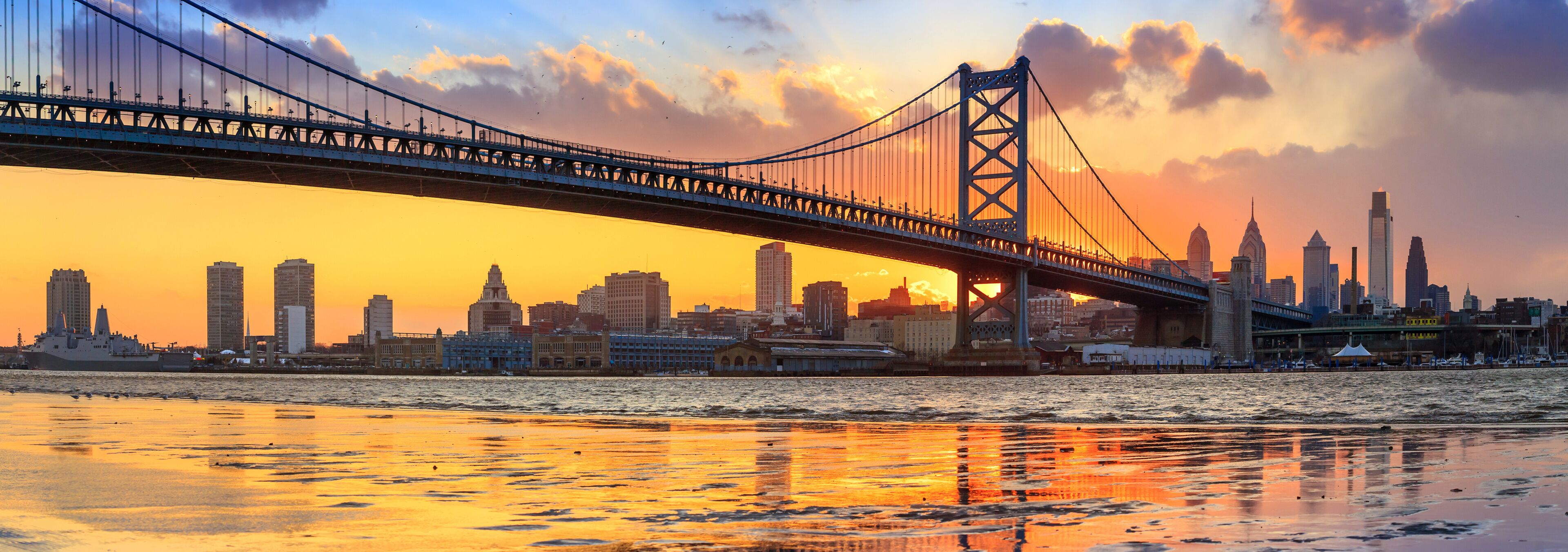 A suspension bridge over a river with a city skyline in the background during sunset.