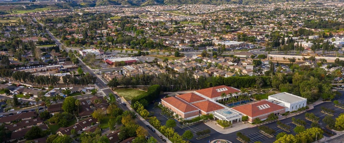 A cityscape with a prominent building complex featuring a red-roofed structure and a white building, surrounded by residential areas and a backdrop of mountains.