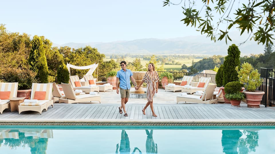 A poolside area with lounge chairs, a couple walking, and a view of distant hills.