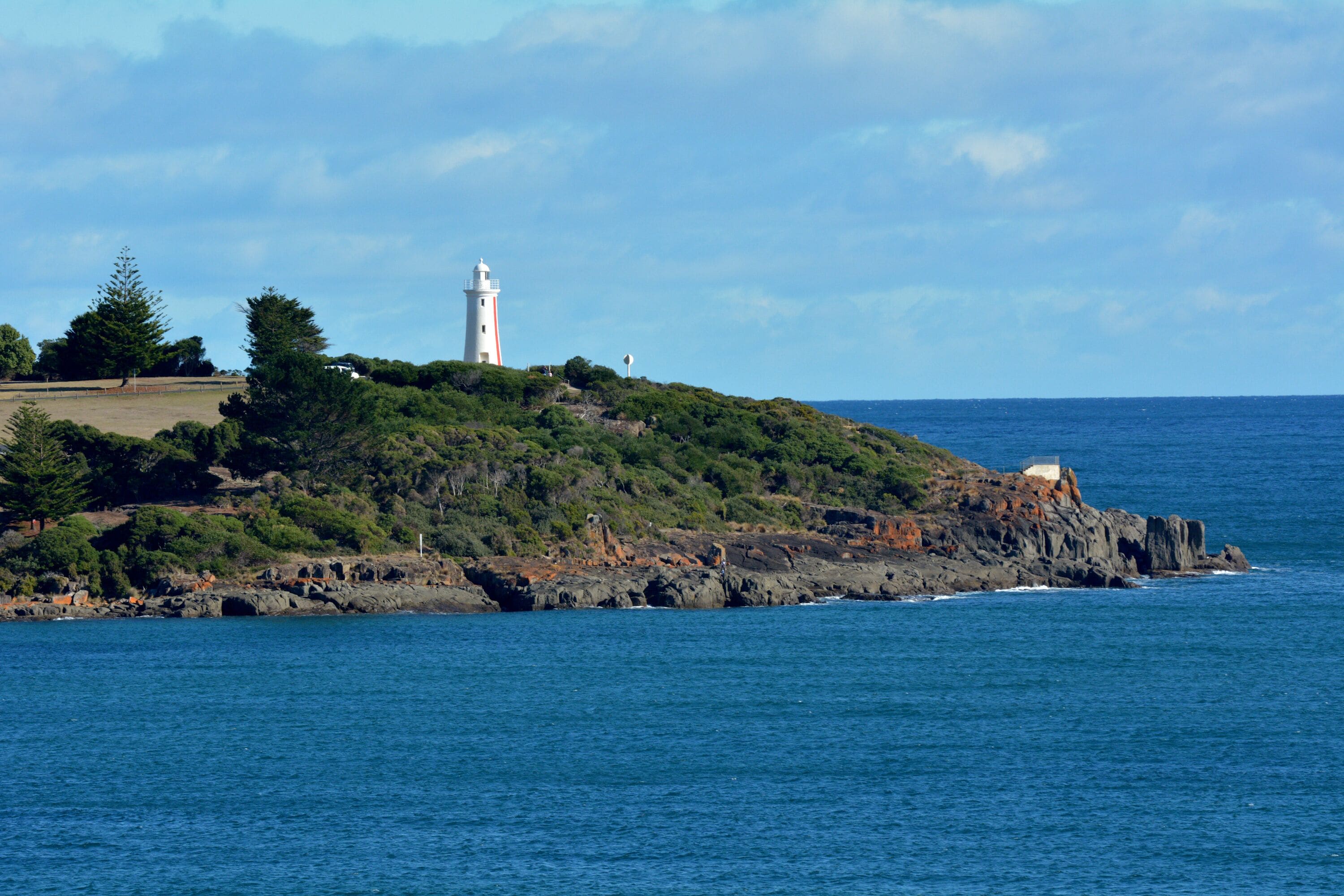 A coastal landscape with a lighthouse on a rocky hill, surrounded by greenery and the ocean.