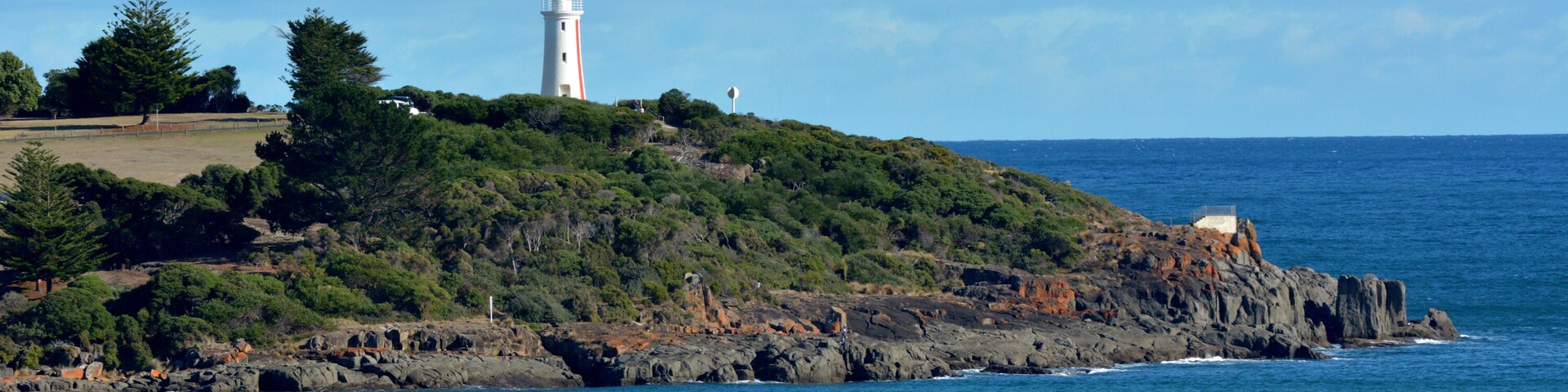 A coastal landscape with a lighthouse on a rocky hill, surrounded by greenery and the ocean.