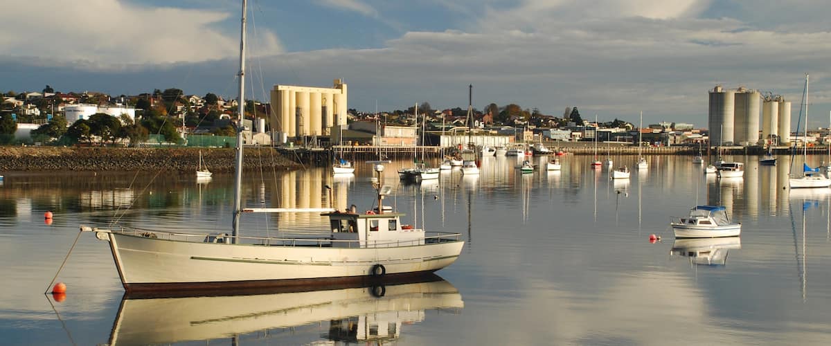 A harbor with several boats docked, a large industrial building, and a calm water body reflecting the sky.