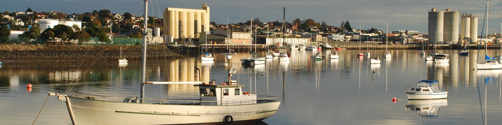 A harbor with several boats docked, a large industrial building, and a calm water body reflecting the sky.