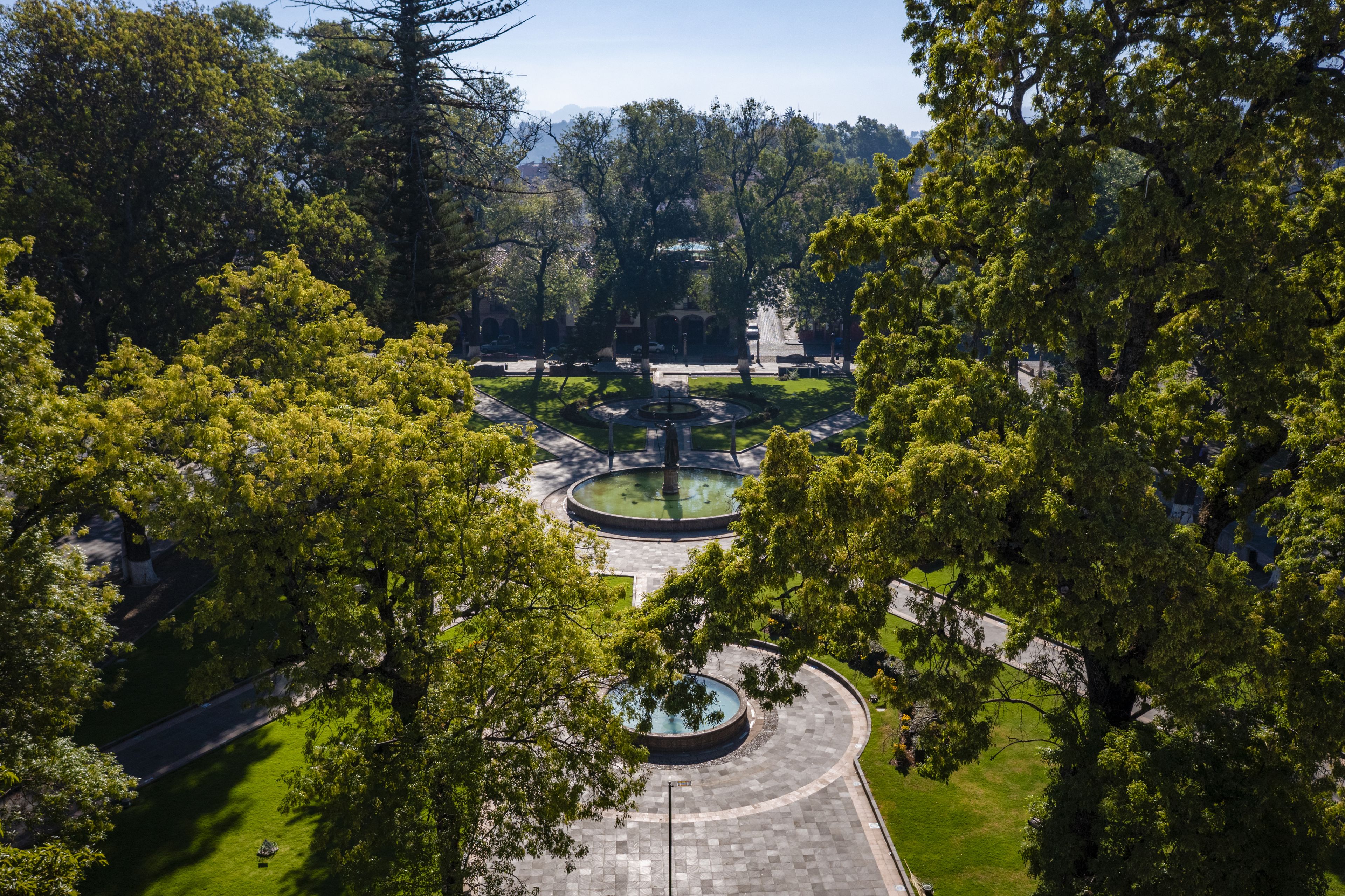 A park with a circular fountain, surrounded by trees and a paved walkway.