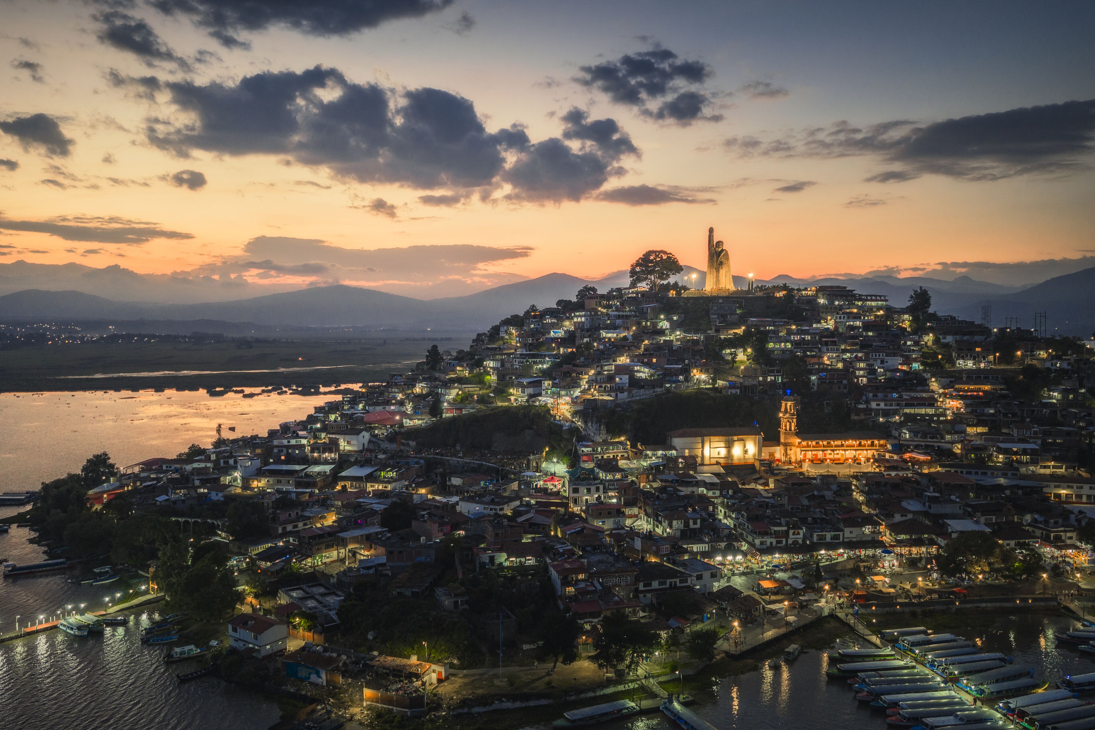 Aerial view of the illuminated statue of Jose Maria Morelos atop Janitzio Island, the vibrant town glowing with warm light against the cool dusk, Janitzio, Michoacan, Mexico.