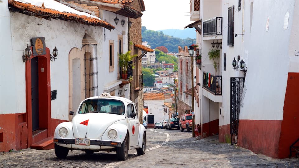 A vintage white taxi driving down a cobblestone street flanked by buildings with red and white facades.