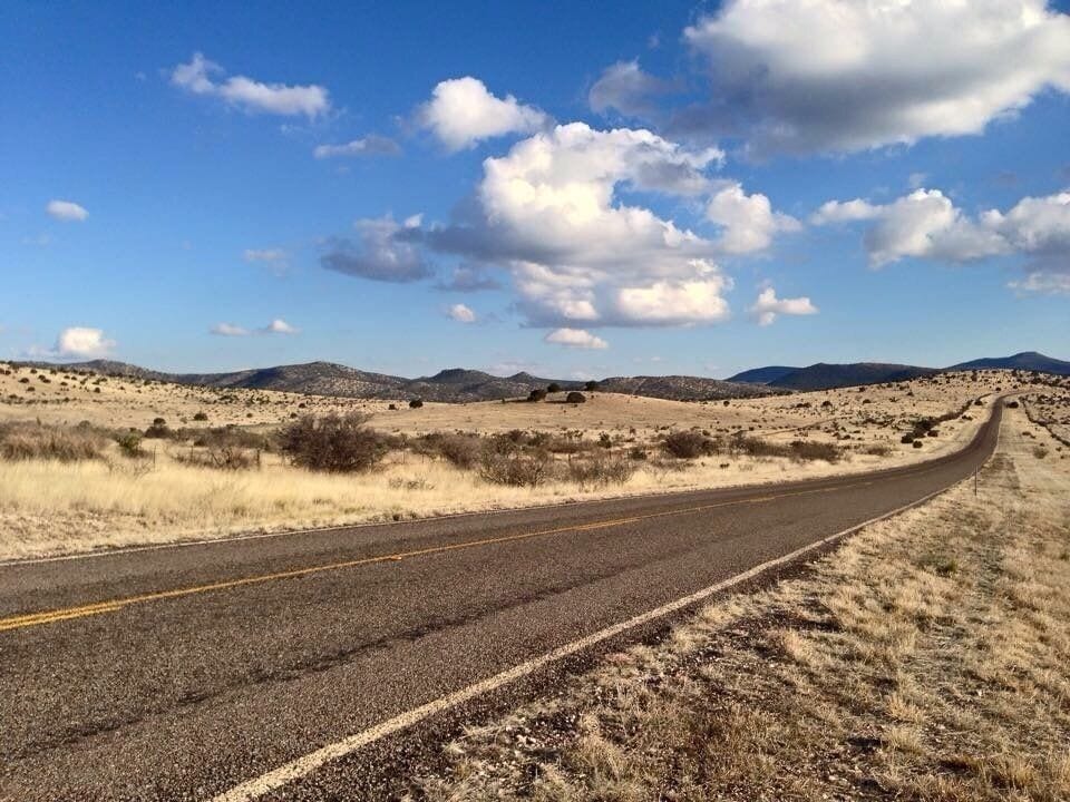 A winding road through a desert landscape with sparse vegetation and distant hills under a partly cloudy sky.