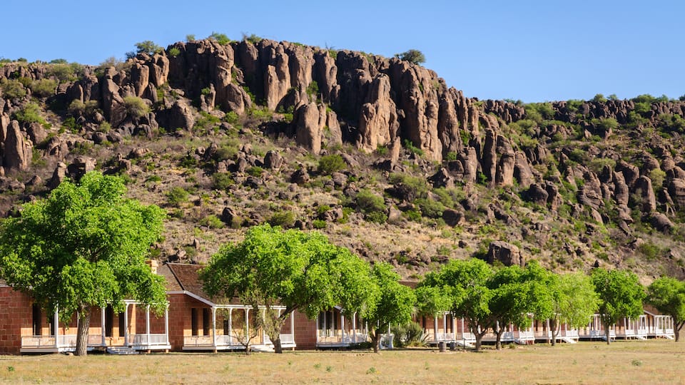 A rocky mountain with a clear sky. In the foreground, there are trees and a building with a porch.