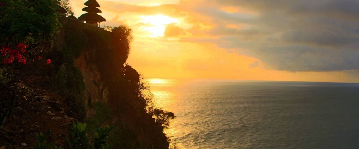A sunset over a coastal cliff with a pagoda and lush greenery.