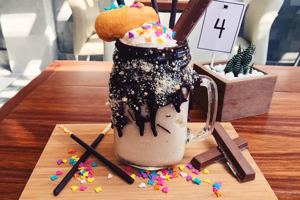 A milkshake in a mug with colorful sprinkles, a donut, and chocolate sticks on a wooden table.