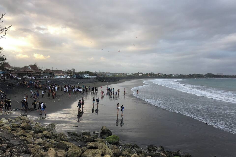 Una playa concurrida, gente caminando por la orilla y un primer plano rocoso.