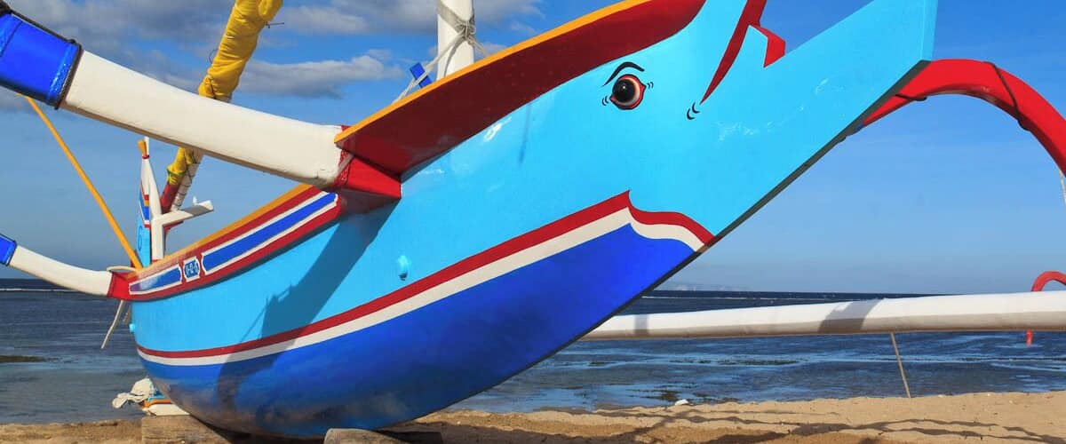 A colorful boat with a blue and red design, resting on a sandy beach with the ocean in the background.