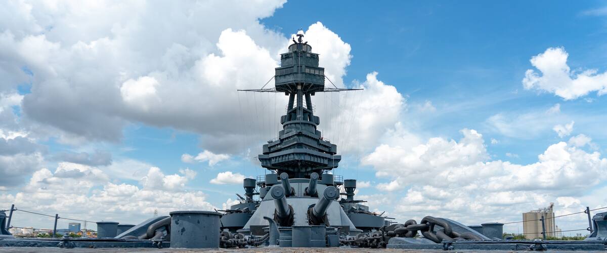 A large naval ship with a central tower and multiple gun turrets, docked on a wooden deck with a clear sky and clouds in the background.