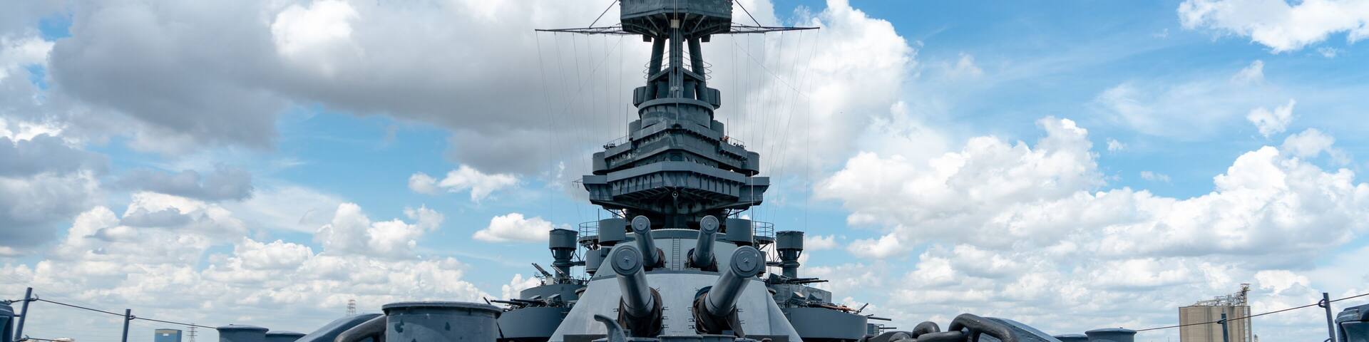 A large naval ship with a central tower and multiple gun turrets, docked on a wooden deck with a clear sky and clouds in the background.