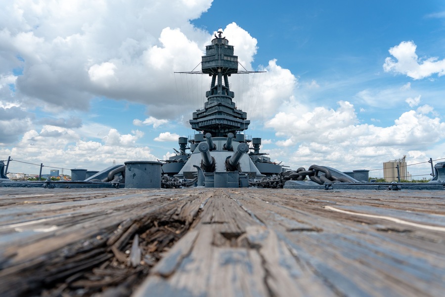 A large naval ship with a central tower and multiple gun turrets, docked on a wooden deck with a clear sky and clouds in the background.