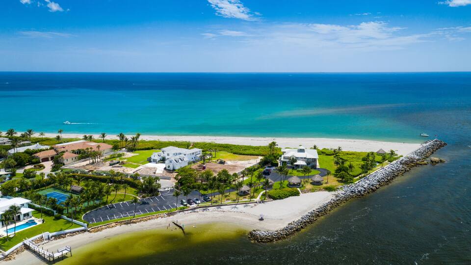 A coastal residential area with a beach, clear blue waters, and a rocky breakwater.