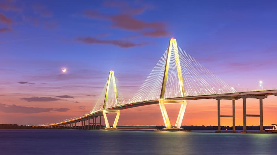 A bridge with a distinctive cable design, spanning over a body of water, with a sunset sky in the background.