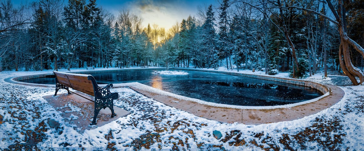 A snow-covered park with a wooden bench, a circular pond, and a forest in the background.