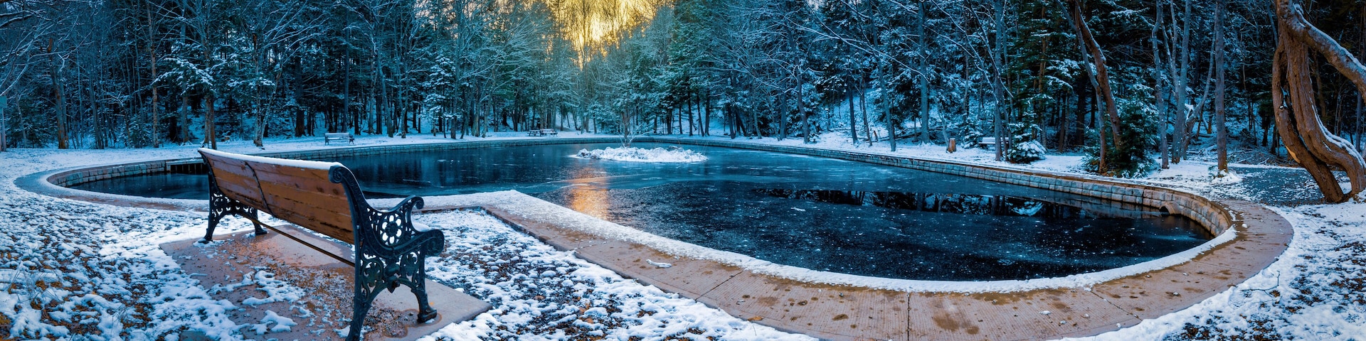A snow-covered park with a wooden bench, a circular pond, and a forest in the background.