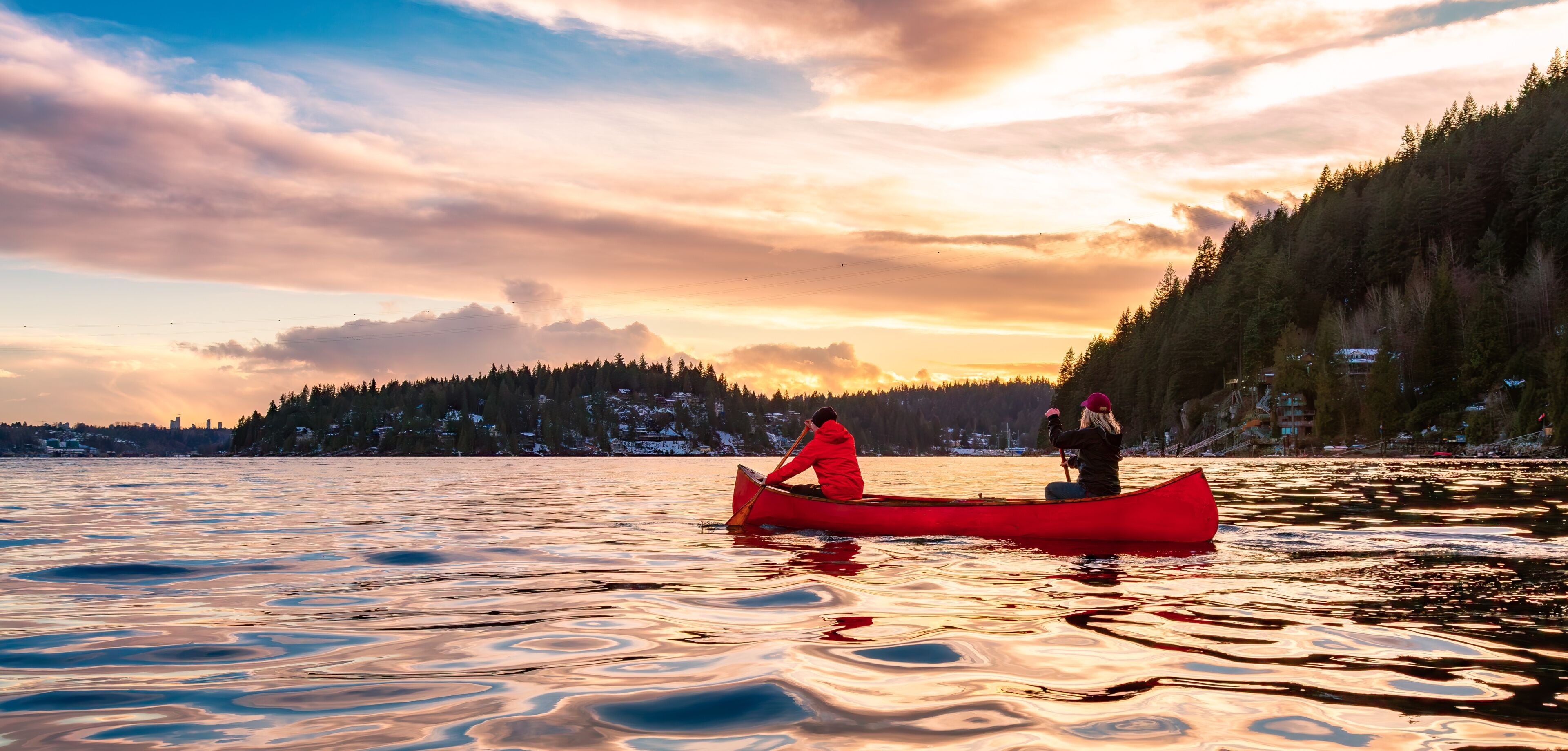 A red canoe on calm water with a sunset sky and a forested hillside in the background.