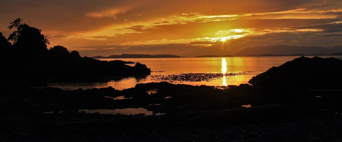A sunset over a rocky shoreline with silhouettes of trees and distant landforms.