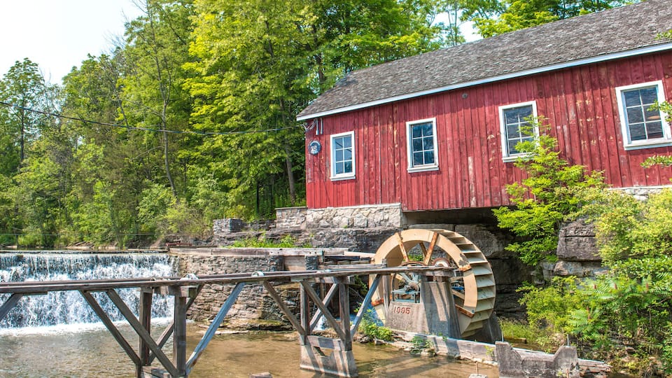 A red wooden mill with a water wheel and a wooden bridge over a river.