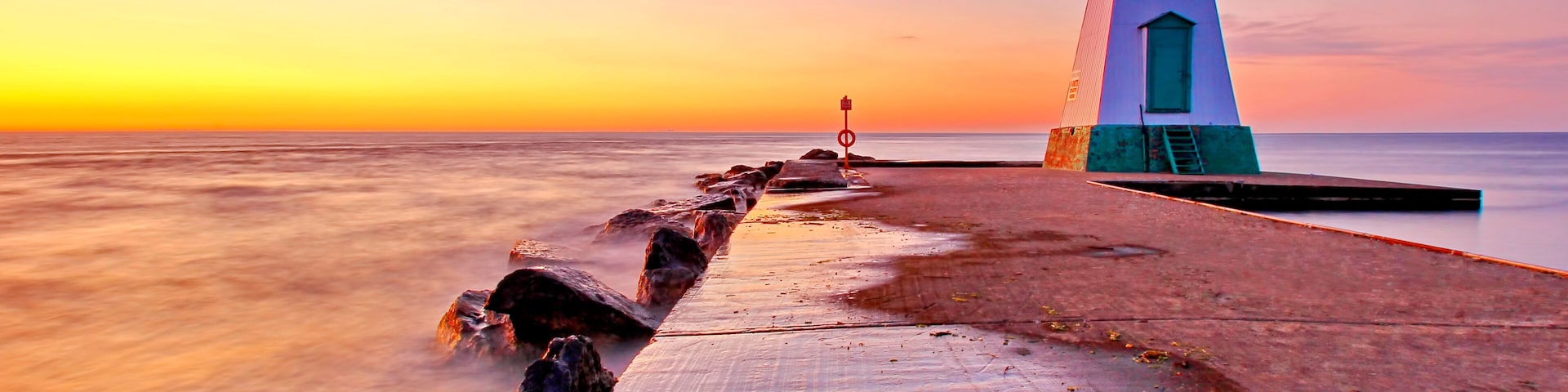 Um farol em um cais com o pôr do sol ao fundo.
