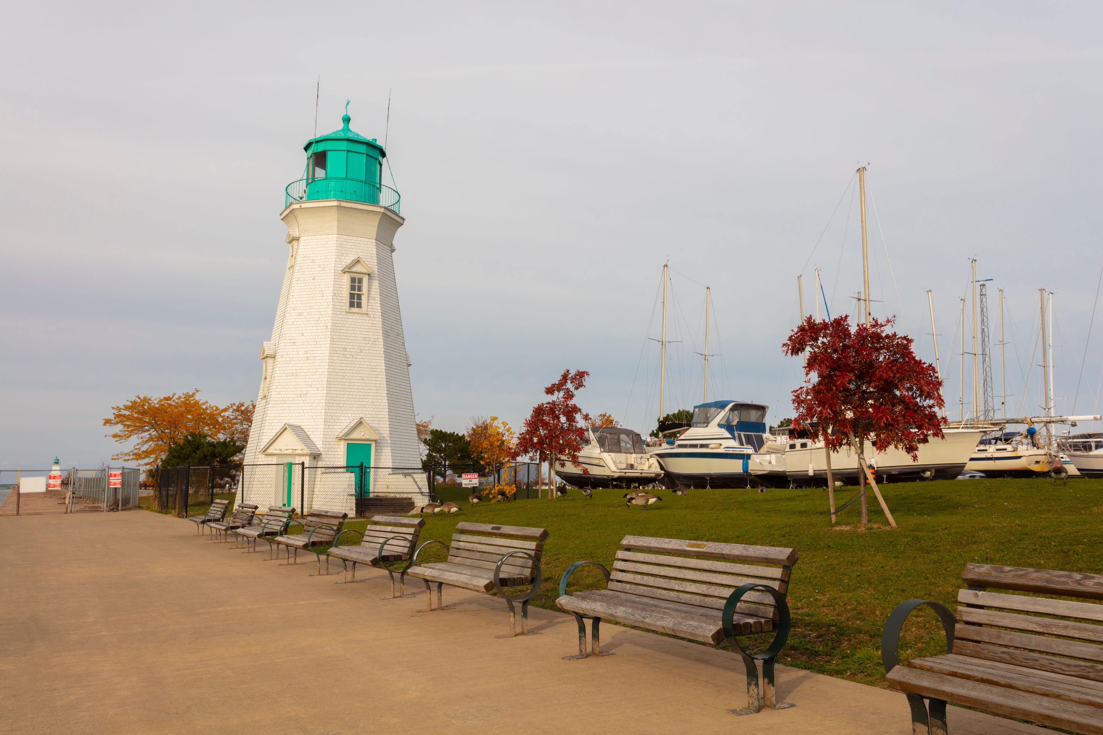 A lighthouse, several benches, a grassy area, and boats docked in the background.