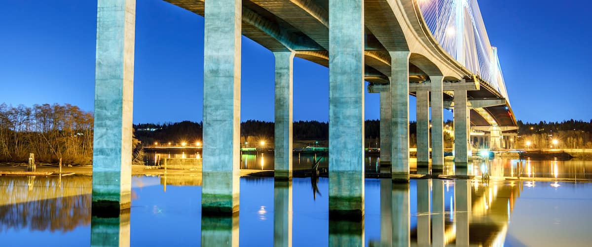 Un pont dont les câbles sont illuminés au crépuscule, surplombant une étendue d’eau calme dans laquelle se reflète la structure du pont.