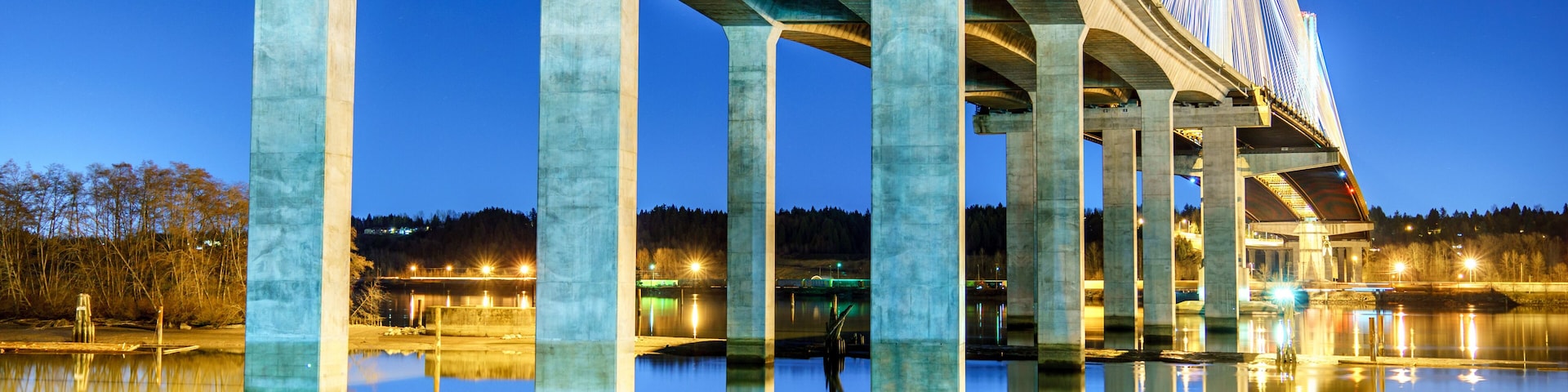 Uma ponte com cabos iluminados ao anoitecer, sobre águas tranquilas que refletem a estrutura.