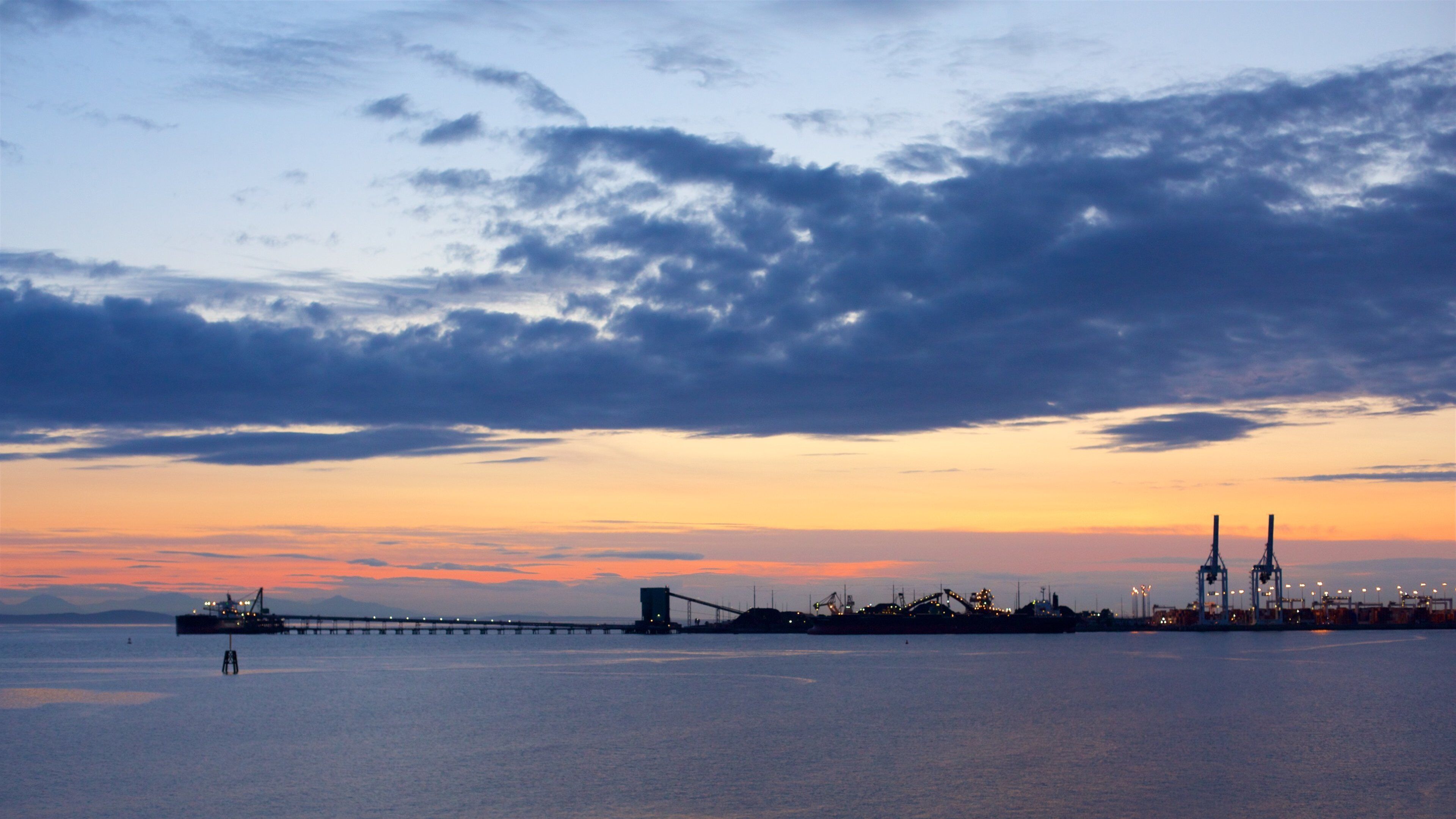A sunset over a body of water with a bridge and industrial structures in the distance.