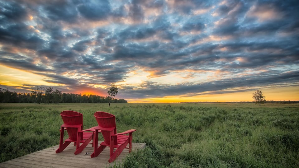 A sunset over a grassy field with three red Adirondack chairs on a wooden deck.