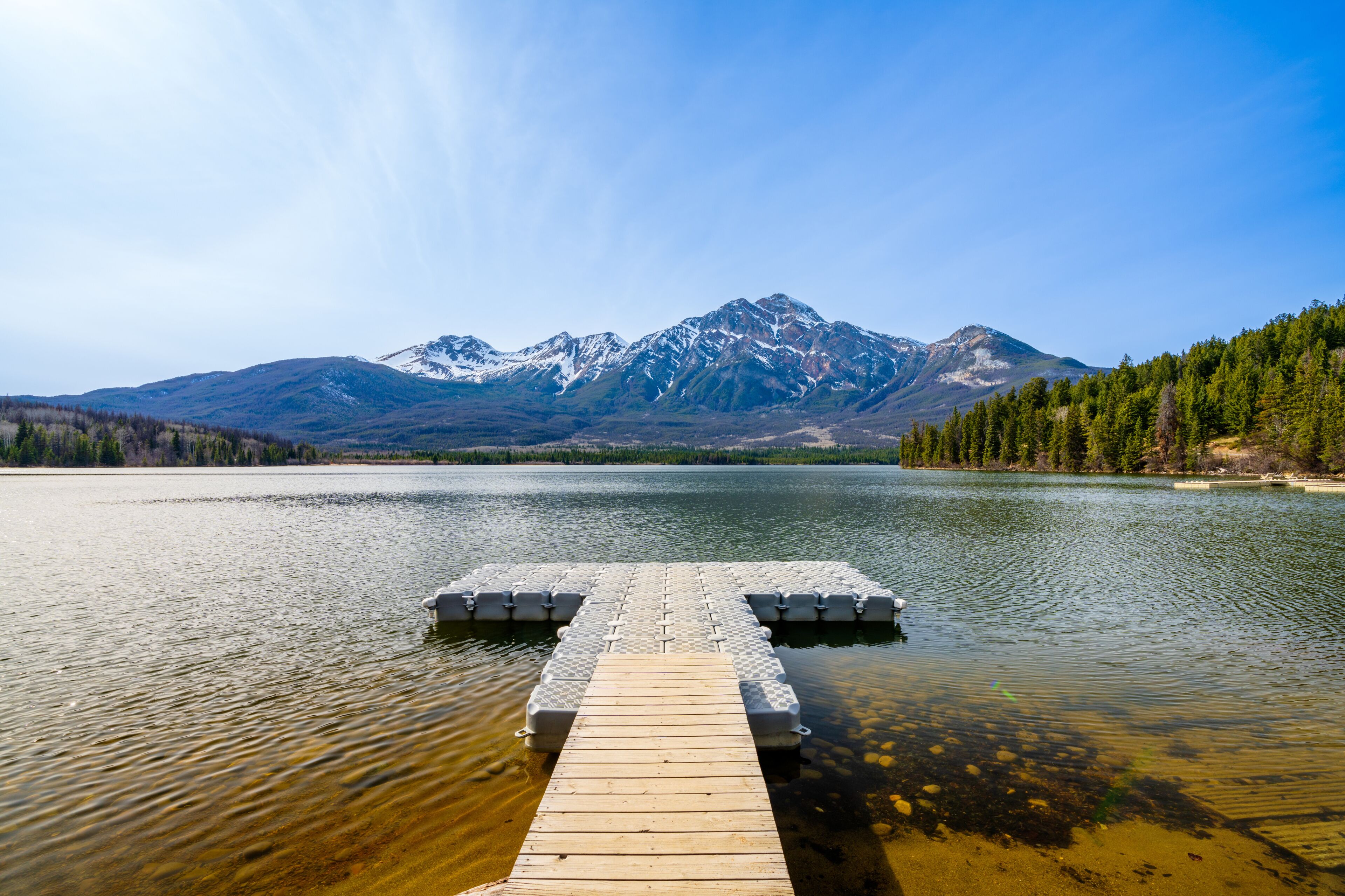 A wooden dock extending into a calm lake with a backdrop of snow-capped mountains and dense forest.