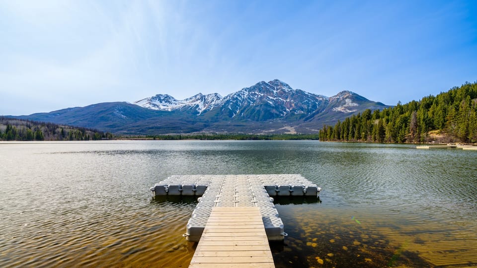 A wooden dock extending into a calm lake with a backdrop of snow-capped mountains and dense forest.