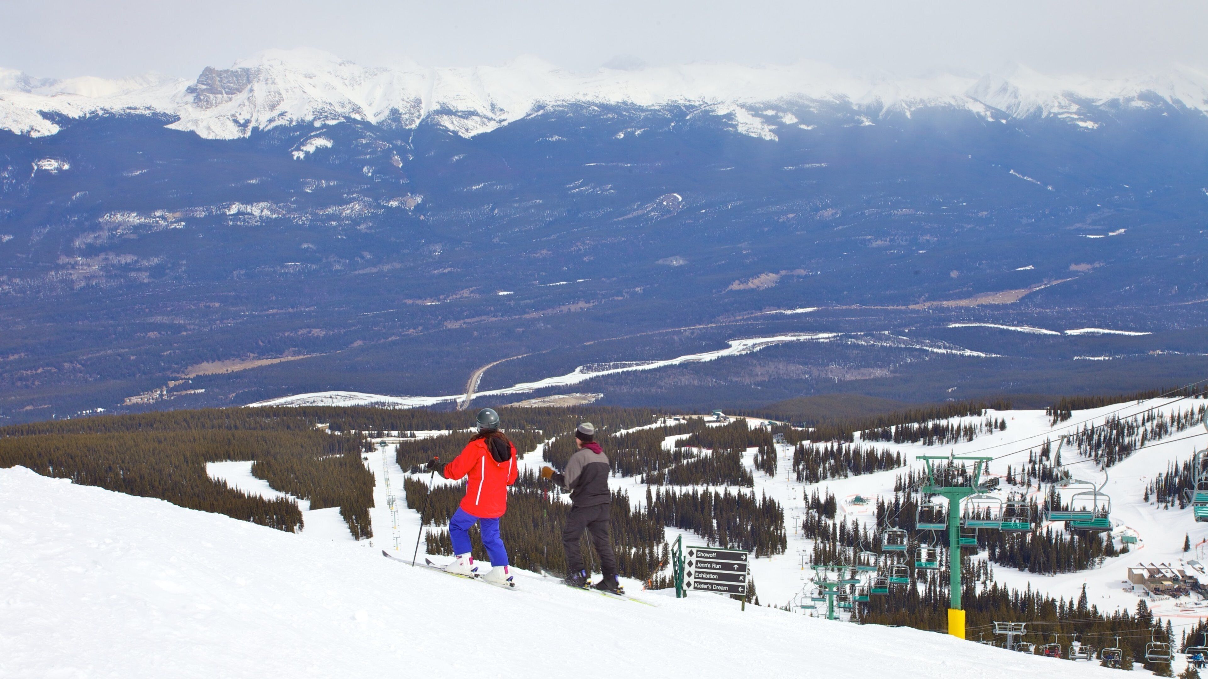 A snowy mountain landscape with skiers, a ski lift, and a distant mountain range.