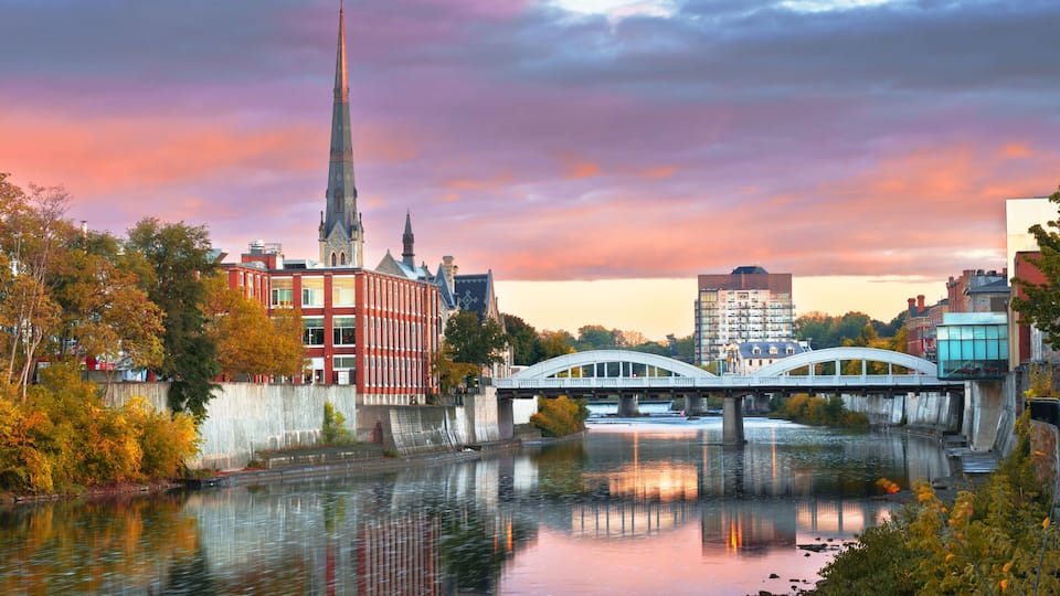 A river with a bridge, a church spire, and autumn-colored trees.