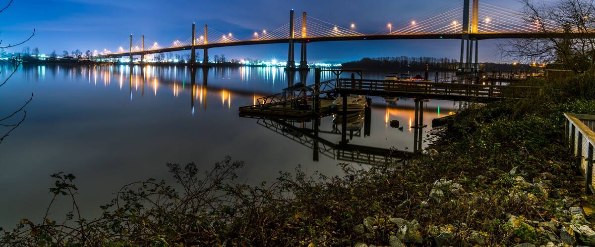 A nighttime view of a suspension bridge over calm water, with city lights reflecting on the surface.