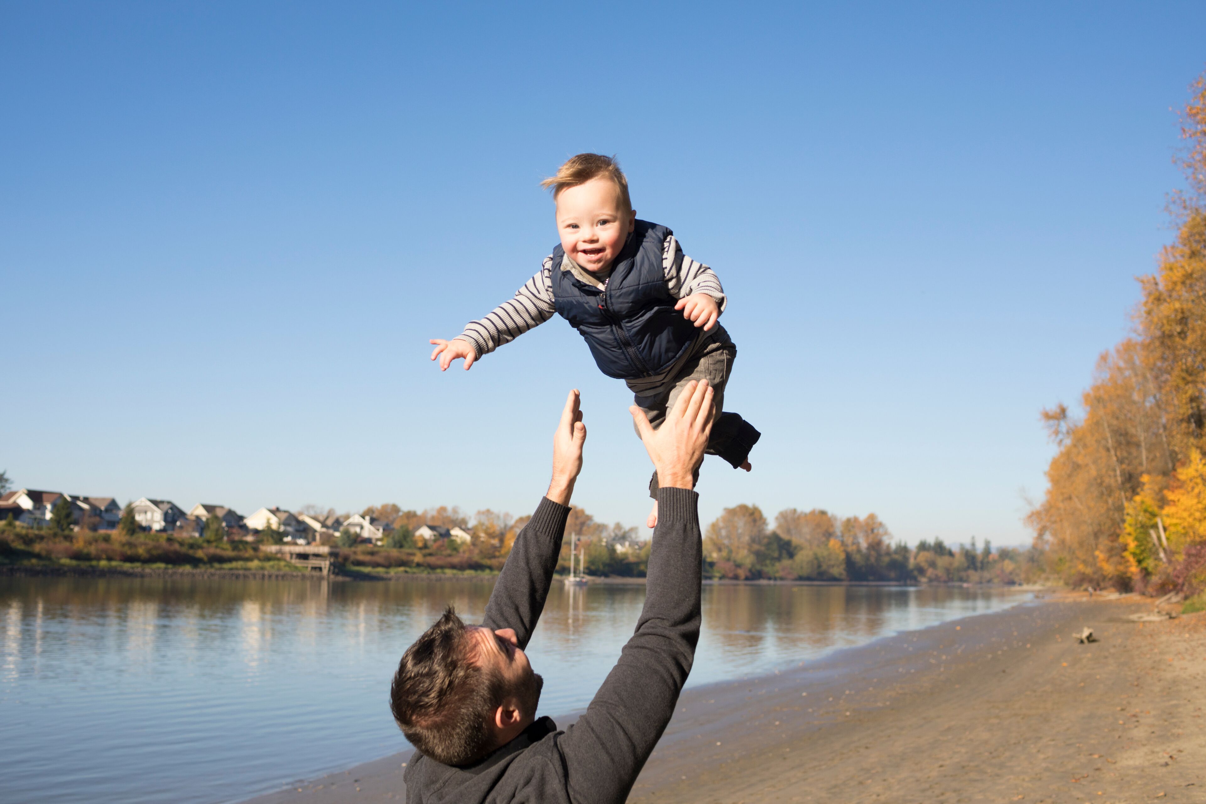 A man lifting a child over a calm lake with houses in the background.