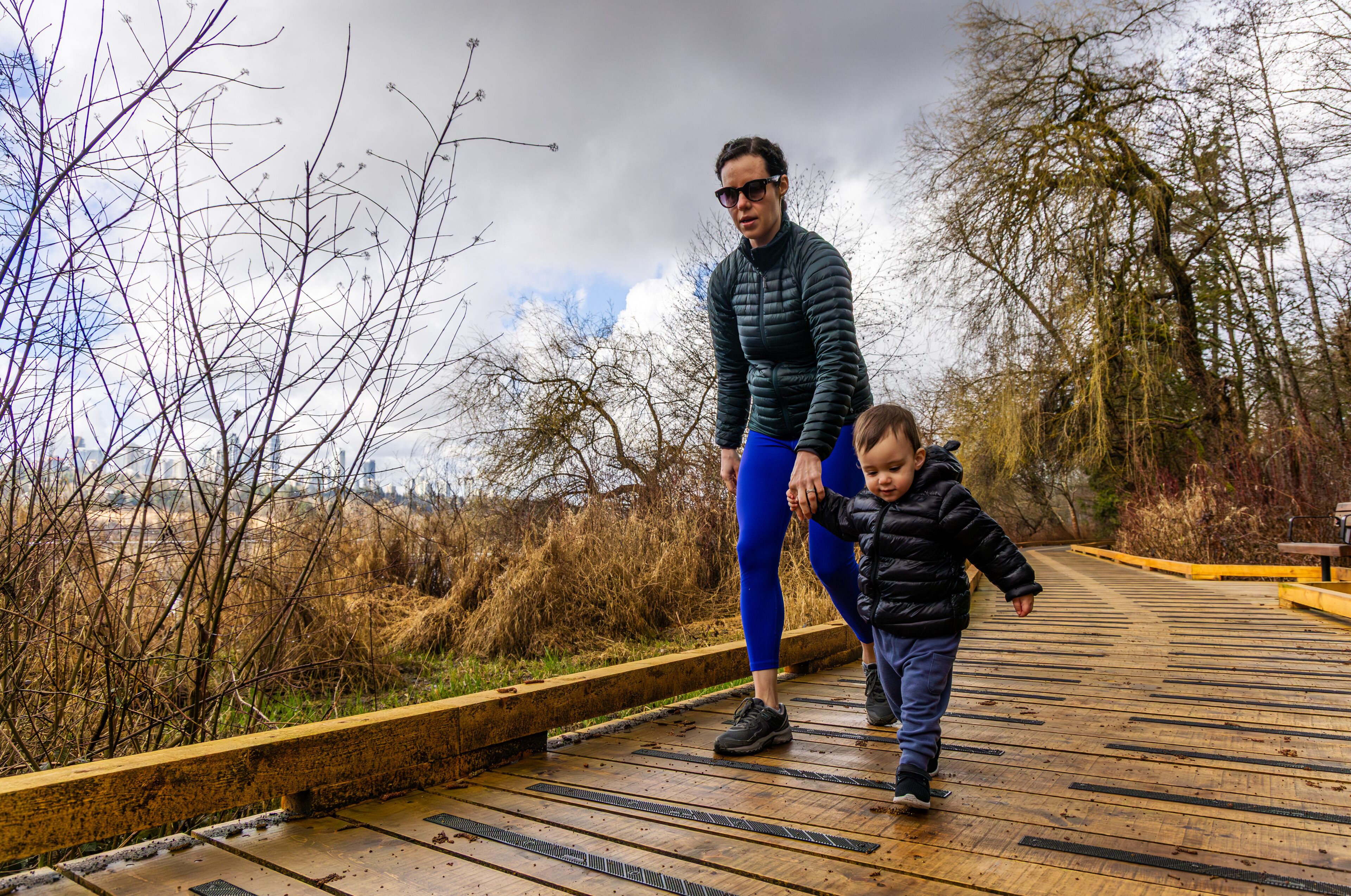 Un paseo marítimo de madera con una persona y un niño caminando, rodeado de vegetación seca y árboles.